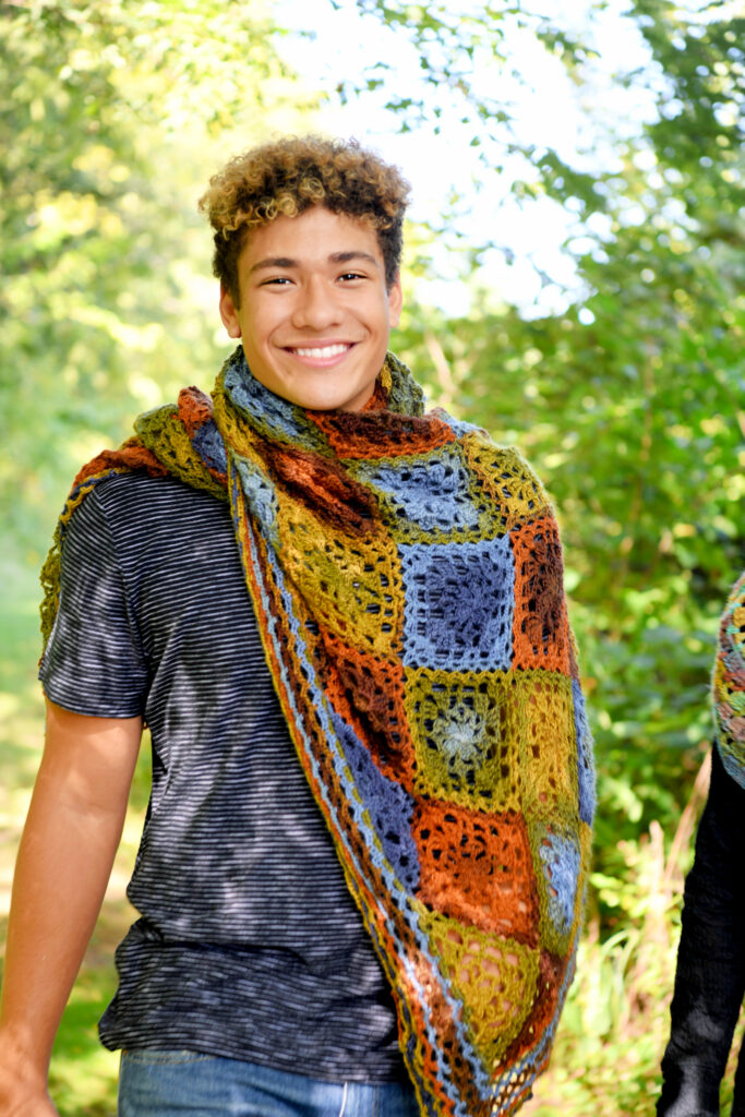 Young man wearing striped tee and blue jeans with shawl wrapped over one shoulder. Background of green leaves and sky. Enchanted Crochet Motif Shawl Free Rectangle Crochet Shawl Pattern - Marly Bird
