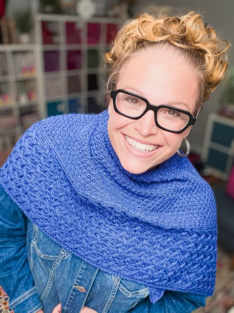 Eowyn cable knit shawl draped over a denim jacket, showing the textured snug cables and double moss stitch in navy blue sport weight yarn.