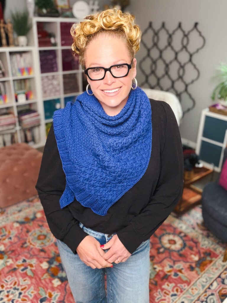 A smiling person models a textured cable knit shawl with visible stitch detail, standing on a colorful rug among books and yarn.