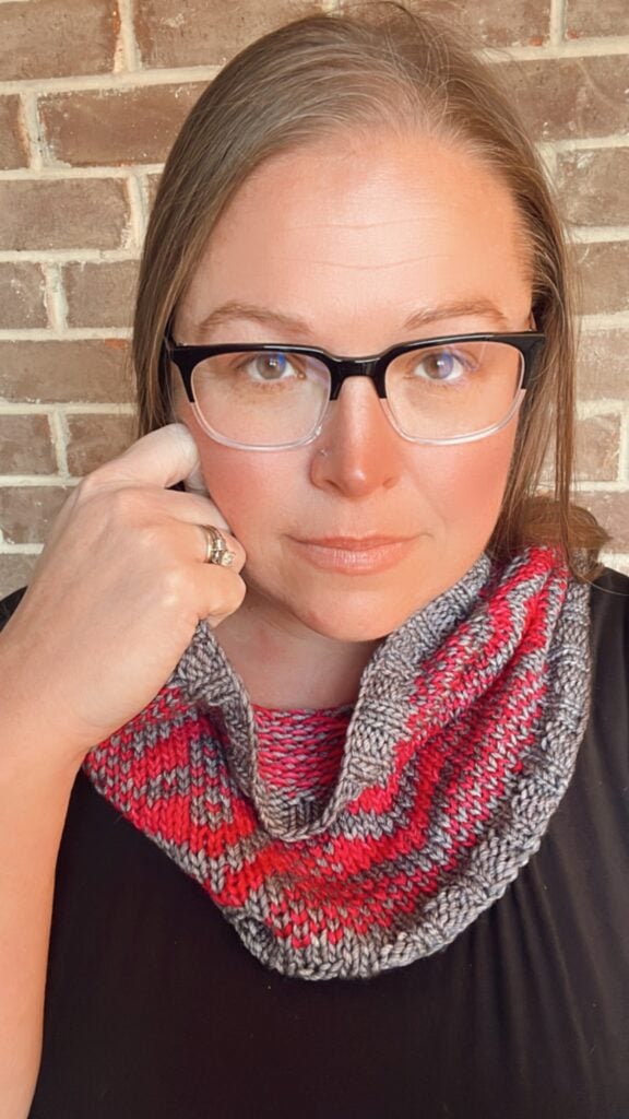 A woman models a hand-knit cowl scarf in red, gray, and pink; the textured stitches are clearly visible.