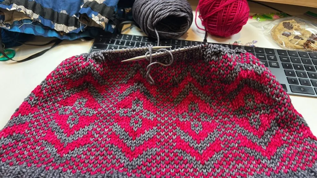 Red and gray stranded knit fabric with diamond motifs on needles, showing neat stitch detail. Yarn balls and cookies in background.