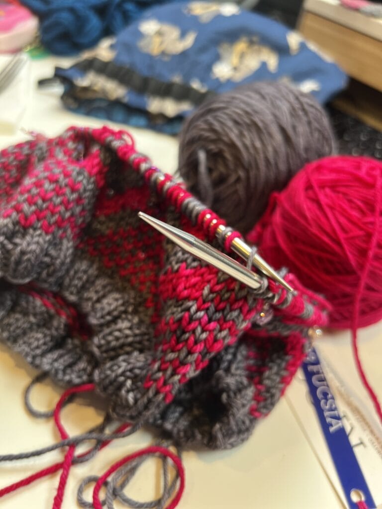 Gray and red knit fabric on metal needles, balls of yarn in background; detailed stitches for a garment project visible.