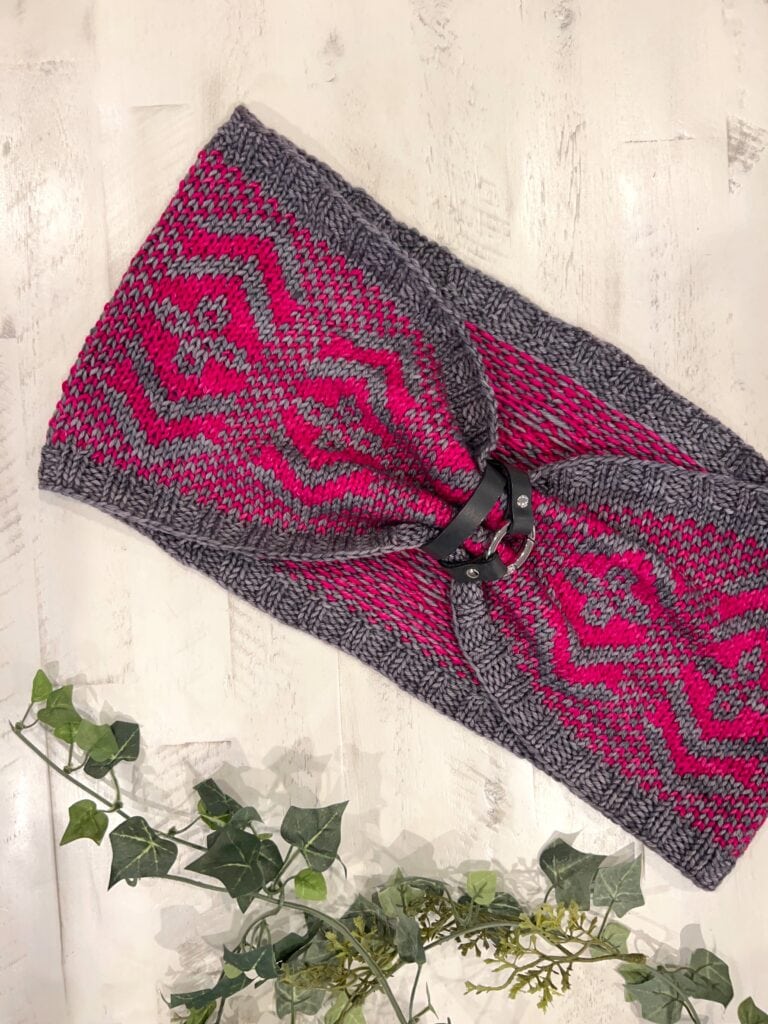 Hand-knit cowl in pink and gray geometric colorwork, folded with black ring; textured stitches visible, displayed by leafy vine.