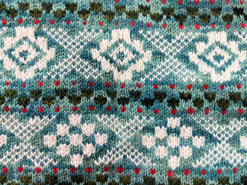 The image displays a detailed view of a knit fabric with stranded colorwork. The pattern features geometric shapes and motifs in a palette of soft whites, blues, greens, and small accents of red, creating a visually rich and textured appearance. This type of work is characteristic of meticulous knitting techniques that involve carrying multiple yarns and alternating between them to create intricate designs. The precision of the stitches and the clarity of the color transitions showcase the skill involved in creating such a piece. The fabric's design is indicative of the complexity and beauty found in stranded colorwork knitting projects like those created by Marly Bird.