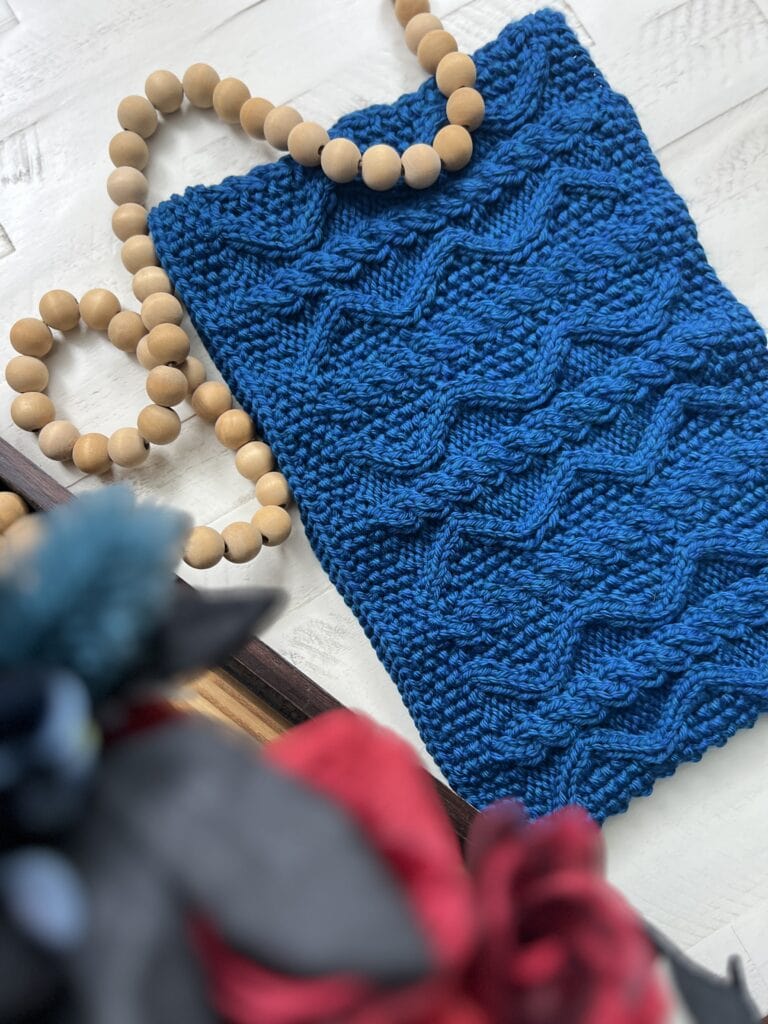 Blue knitted bag with zigzag texture and round wooden bead handle on white surface, flowers blurred in foreground.