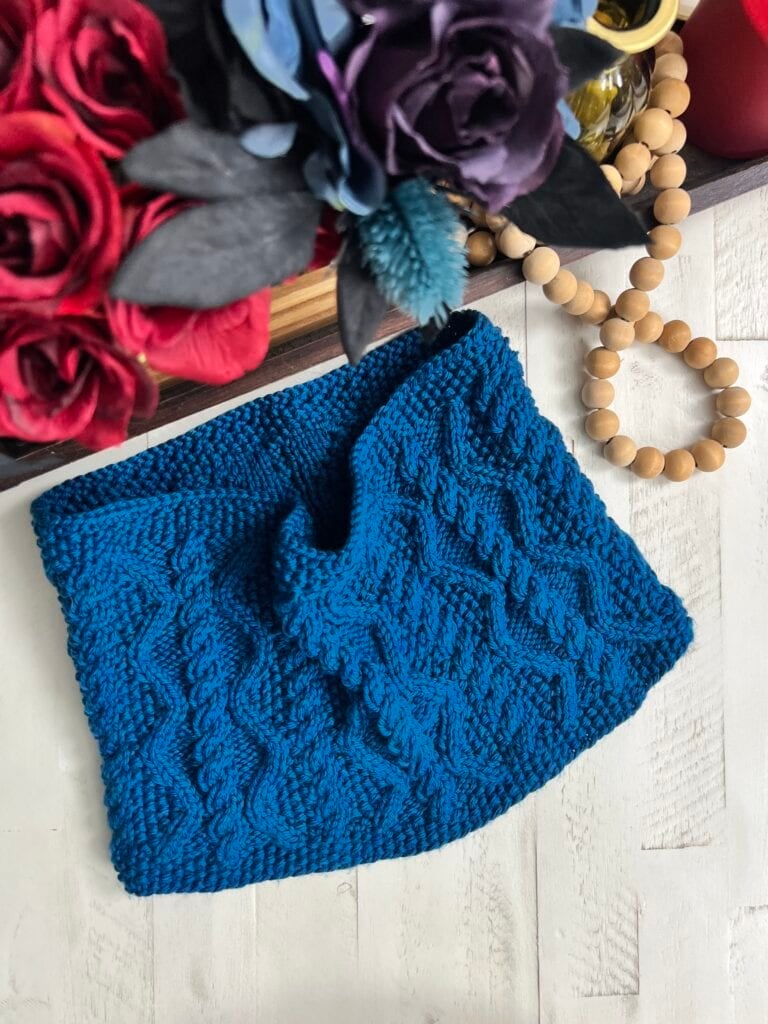 Blue cable knit cowl with wavy texture displayed on white wood, accented by a beaded bracelet and fabric bouquet.