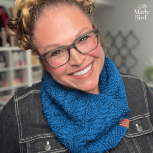 Person models a textured blue cable knit cowl, showing detailed stitches; shelves of yarn and tools are softly blurred behind.