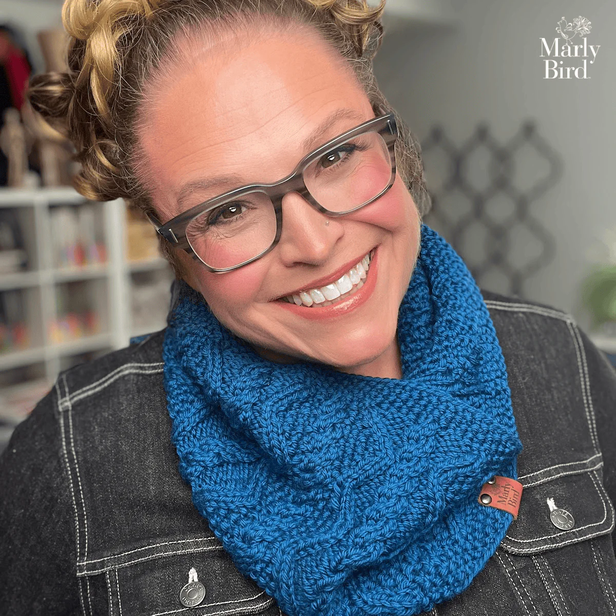 Person models a textured blue cable knit cowl, showing detailed stitches; shelves of yarn and tools are softly blurred behind.
