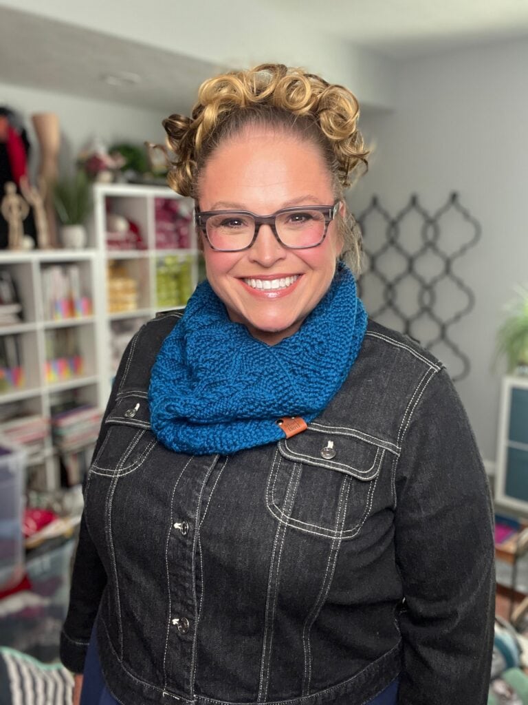 Woman wears a blue cable knit cowl with textured stitch detail, standing among colorful fabrics and craft supplies in a bright room.