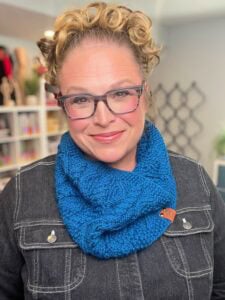 A person models a blue cable knit cowl, showing off its textured stitches; they wear glasses and a dark denim jacket.