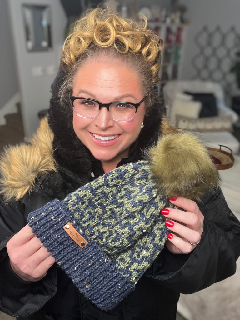 A woman with glasses and curly hair smiles while holding a Falling Leaves Mosaic Knit Hat with a fur pom-pom. She is wearing a winter coat with a fur trim, and the cozy living room background enhances her joy at discovering the free pattern's mosaic tips. -Marly Bird