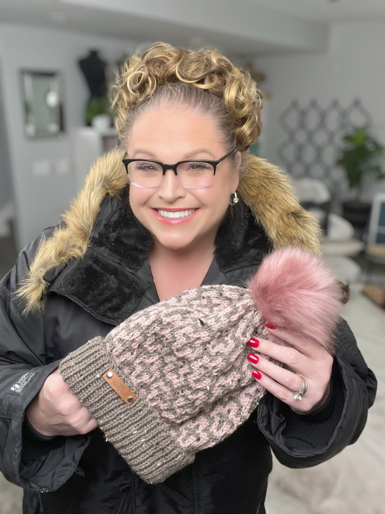 A woman with curly hair and glasses is smiling, holding a Falling Leaves Mosaic Knit Hat adorned with a pink pom-pom. She's wearing a black coat with a fur-trimmed hood. The background reveals a cozy room with a plant and some furniture, offering hints of mosaic tips for home decor enthusiasts. -Marly Bird