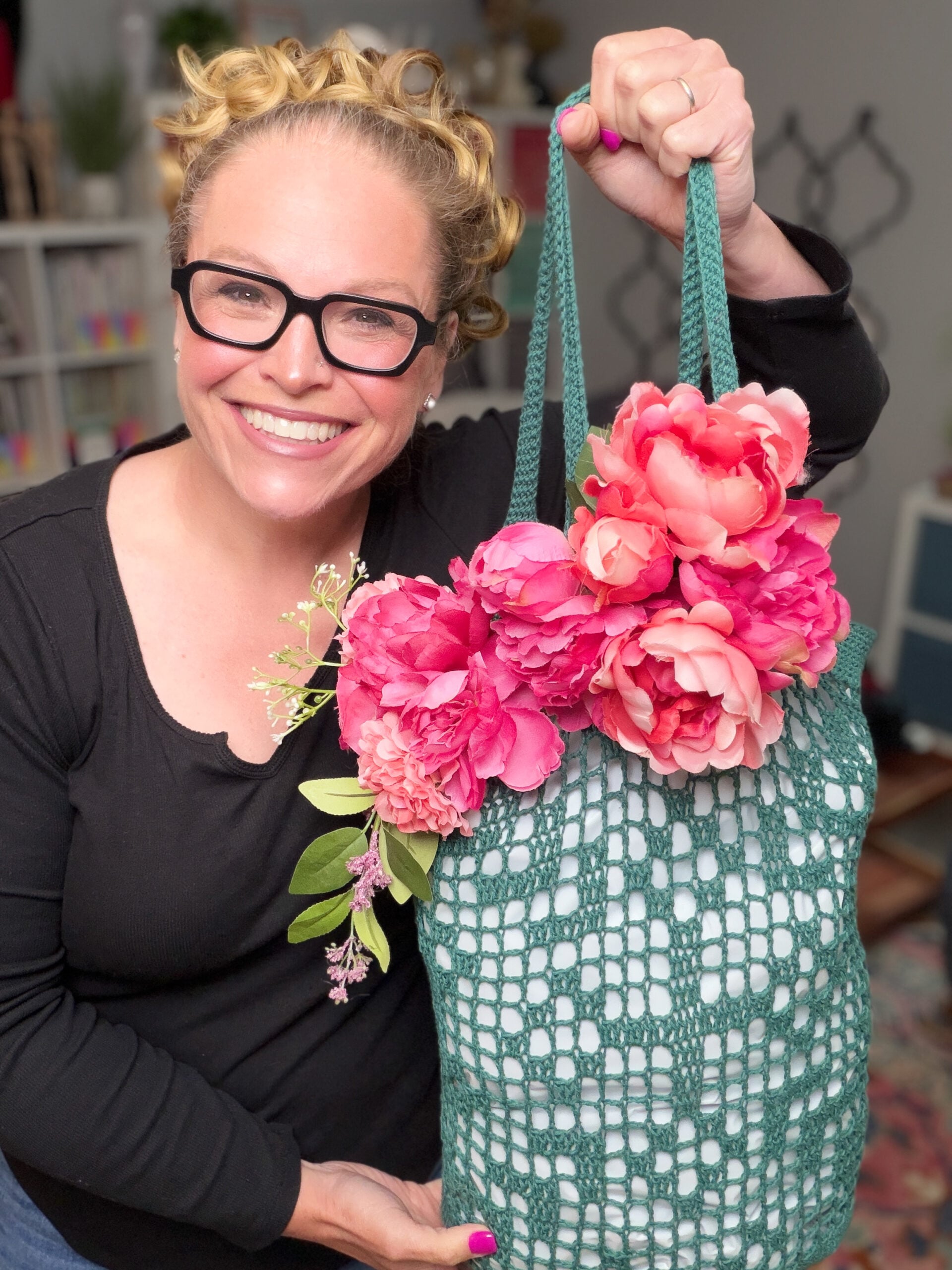 Green filet crochet bag with large pink artificial flowers, shown by its maker indoors; visible stitch detail and soft texture.