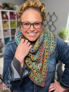 Person models a colorful textured crocheted triangle shawl over a denim jacket, with yarn shelves behind them.