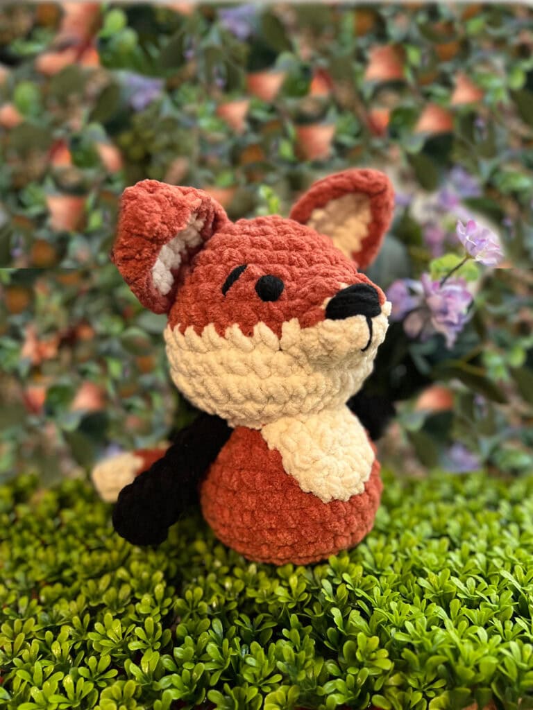 Amigurumi fox plush made with orange, white, and black yarn; sits on moss with visible stitches and soft texture.