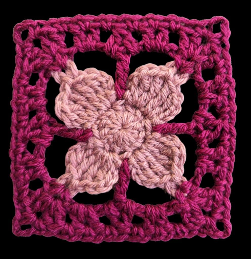 A crocheted granny square with a four-petal flower in the center—light purple petals framed by a dark magenta square on a black background. This vibrant crochet motif is perfect inspiration for your next Crochet Motif Shawl Pattern or Fernanda Shawl. -Marly Bird