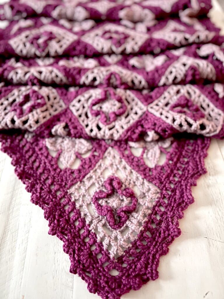 Close-up of a crocheted blanket featuring intricate diamond-shaped patterns in shades of purple and lavender, with lacy, textured designs, laid out on a white surface. -Marly Bird