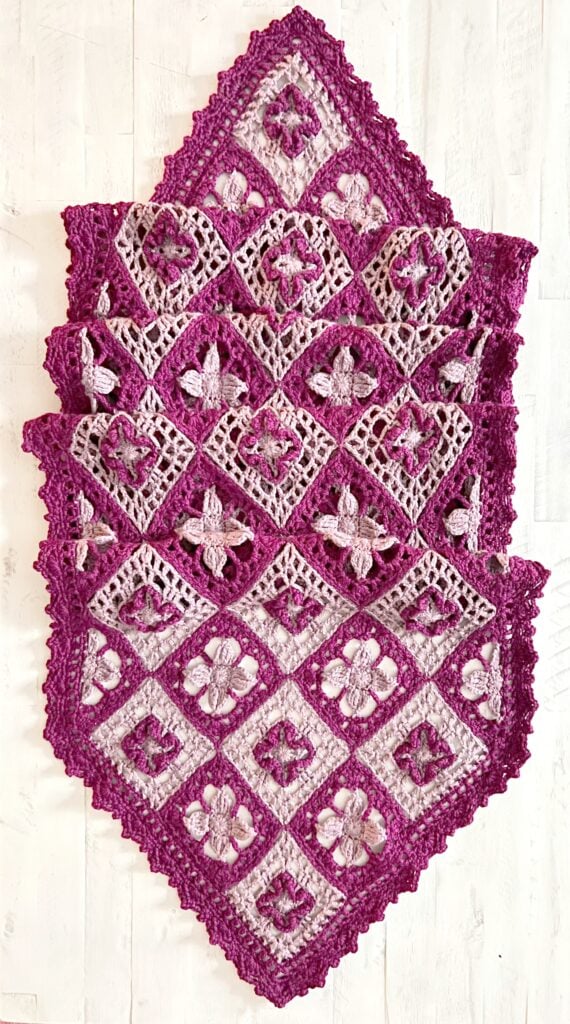 A handmade crochet shawl with a diamond-shaped pattern, featuring purple and lavender floral motifs joined together, displayed on a white wooden surface. -Marly Bird