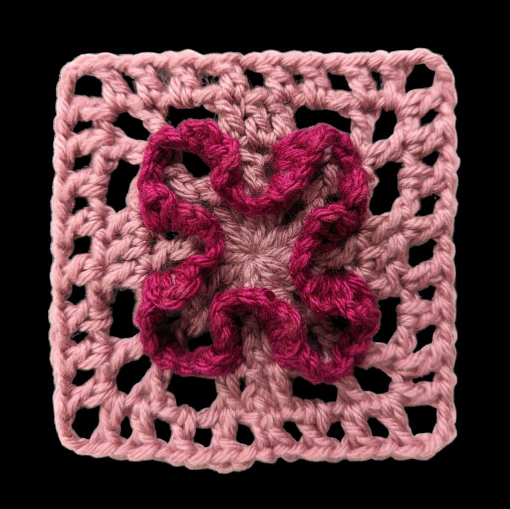 A pink crocheted granny square with an intricate, raised, dark pink flower-like pattern in the center, inspired by the Marly Bird Crochet Motif Shawl Pattern, set against a black background. -Marly Bird