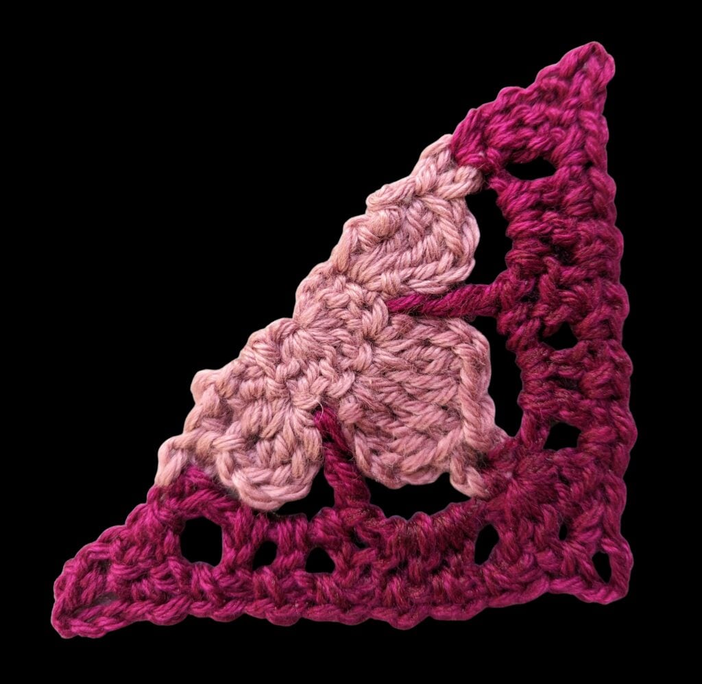 A crocheted triangle motif inspired by the Crochet Motif Shawl Pattern, featuring two pink butterfly wings in the center with gradient yarn in shades of pink and burgundy, displayed against a black background. -Marly Bird