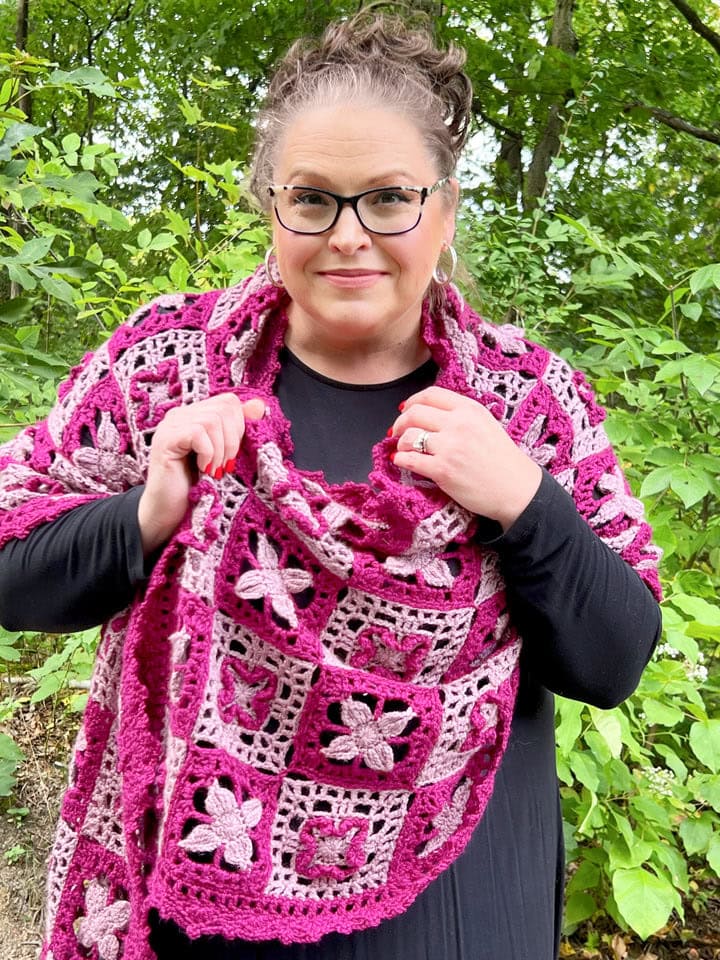 A woman with glasses and curly hair pulled up wears a black dress and drapes a pink and magenta shawl, made from free knitting and crochet patterns with floral square motifs, around her shoulders, standing outdoors in front of green foliage. -Marly Bird