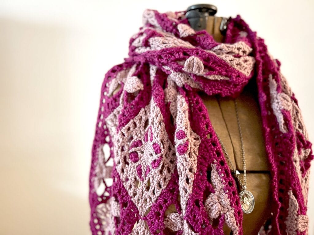 A mannequin displays a crocheted shawl in shades of pink and purple with intricate square patterns, inspired by free knitting and crochet patterns. The mannequin also wears a long necklace with a round pendant. The background is softly blurred. -Marly Bird