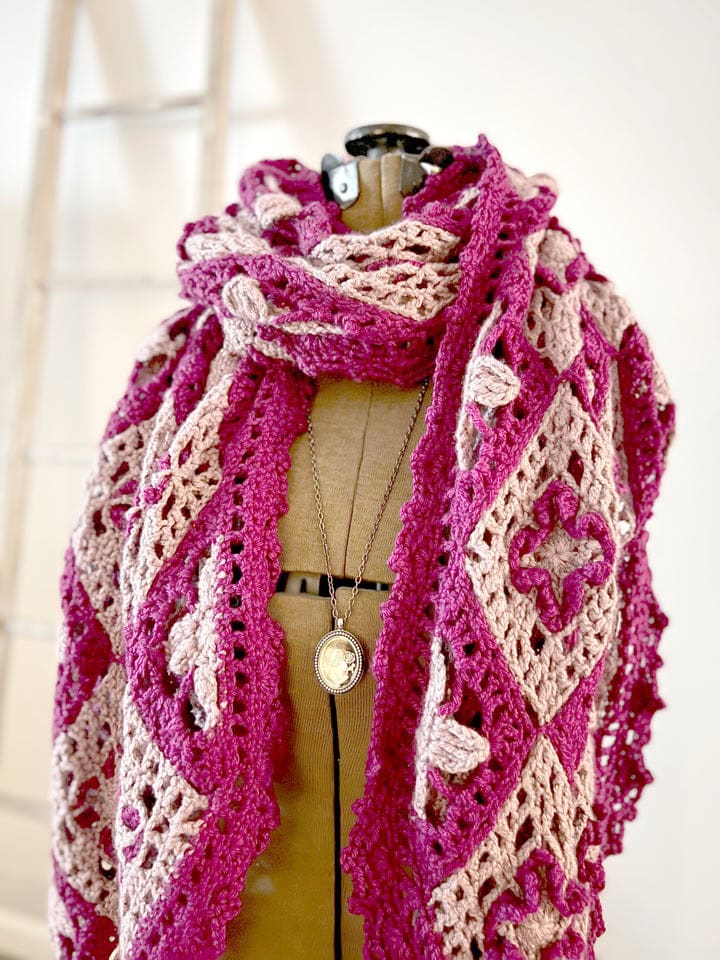 A dress form displays a chunky, crocheted scarf in pink and beige with intricate floral and geometric patterns, perfect inspiration for free knitting and crochet patterns. The dress form also wears a long necklace; a wooden ladder stands in the background. -Marly Bird