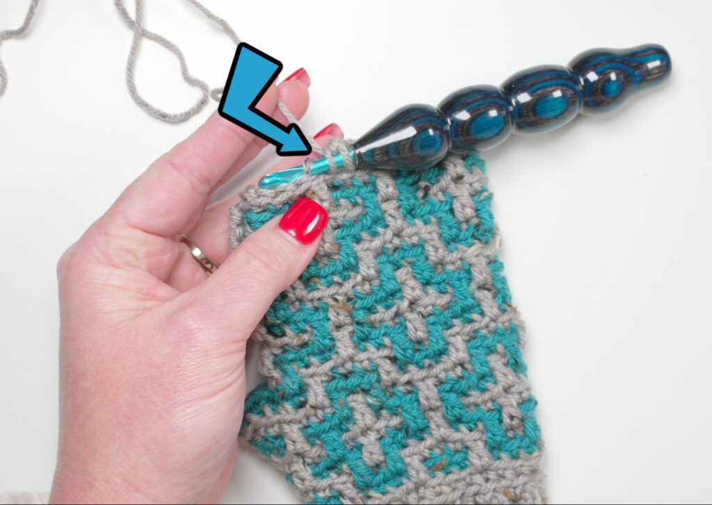 Close-up of a hand crocheting a textured pattern for fingerless mittens using blue and gray yarn. The hand holds a unique, smooth, glass-like blue crochet hook. A blue arrow highlights the stitches, emphasizing the technique from the free pattern "Cinnamon Dreams. -Marly Bird