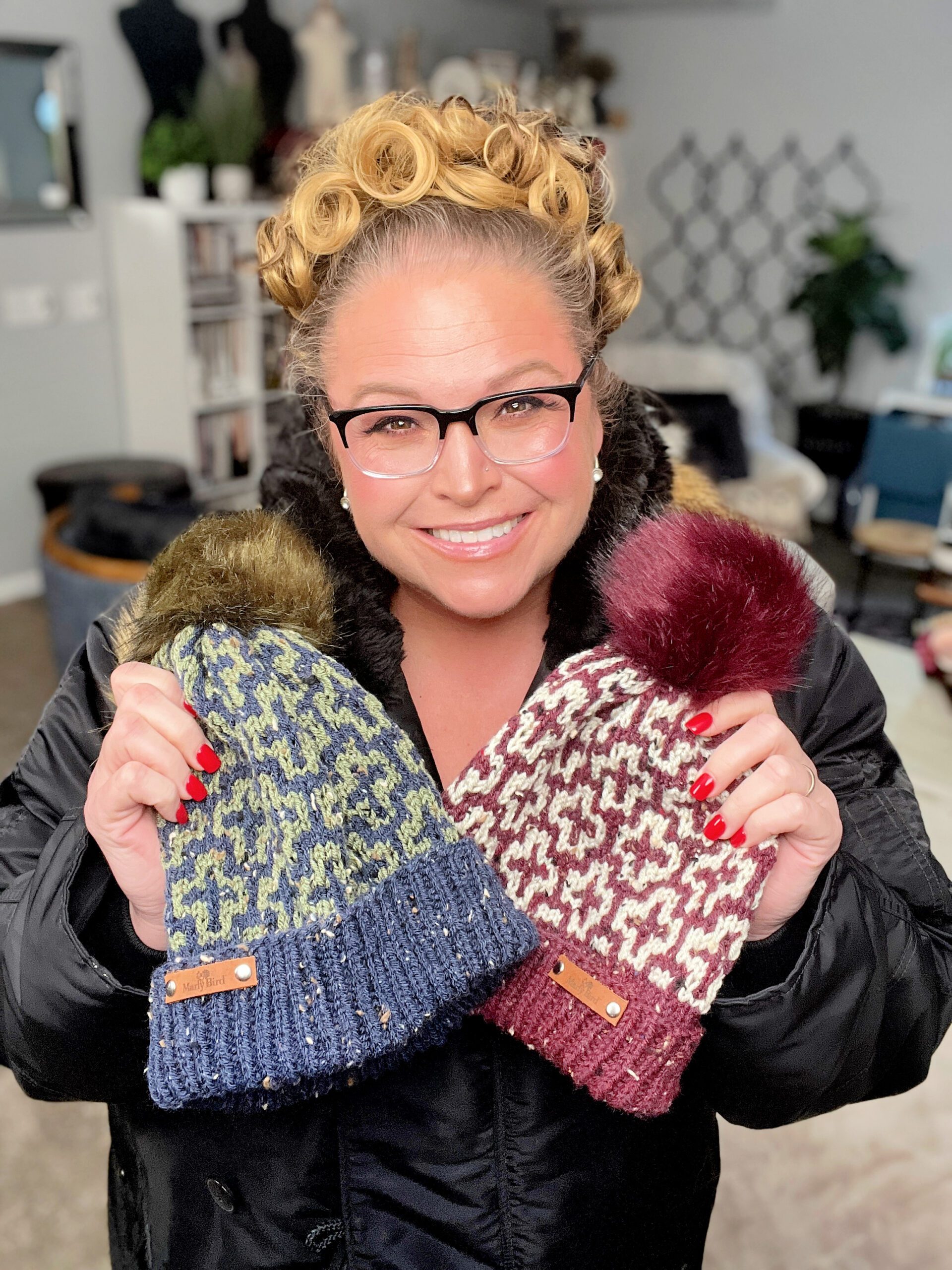 A person wearing glasses smiles while holding two knitted hats with pom-poms, showcasing a free pattern. One hat is blue and green, and the other is red and white. The cozy indoor setting adds warmth with its furnishings and decorations. -Marly Bird