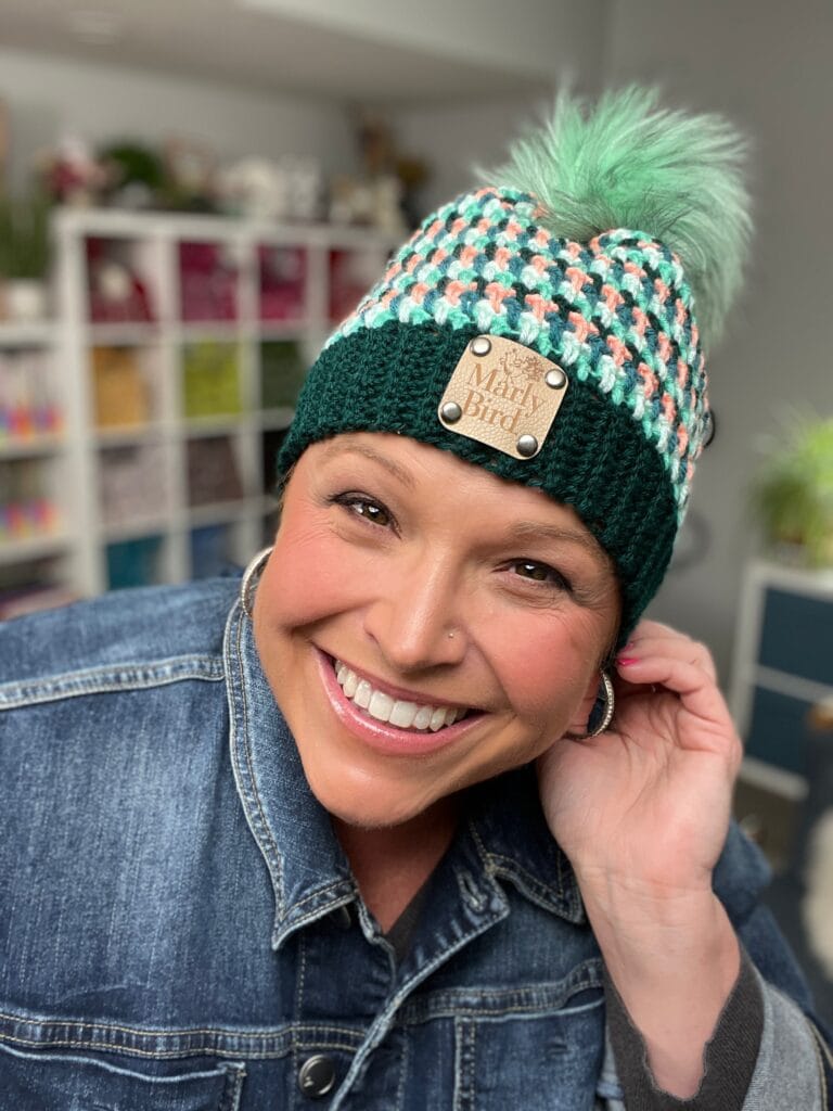 A smiling person wears a green and white patterned knit hat with a pom-pom and “Marly Bird” label—perfect for stash-busting or finding free knit hat patterns—plus a denim jacket and hoop earrings, sitting indoors with shelves and plants behind them. -Marly Bird