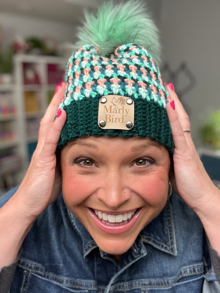 A smiling person in a denim jacket holds a colorful, stash-busting crocheted hat with a pom-pom and a label that says "Marly Bird." The softly blurred background reveals shelves and plants. -Marly Bird