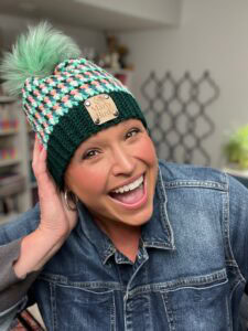 A smiling woman in a green and white knit hat with a pom-pom and a "Yarny Bird" label poses cheerfully indoors, wearing a denim jacket—her cozy look is perfect for showing off stash-busting knit hat patterns. -Marly Bird