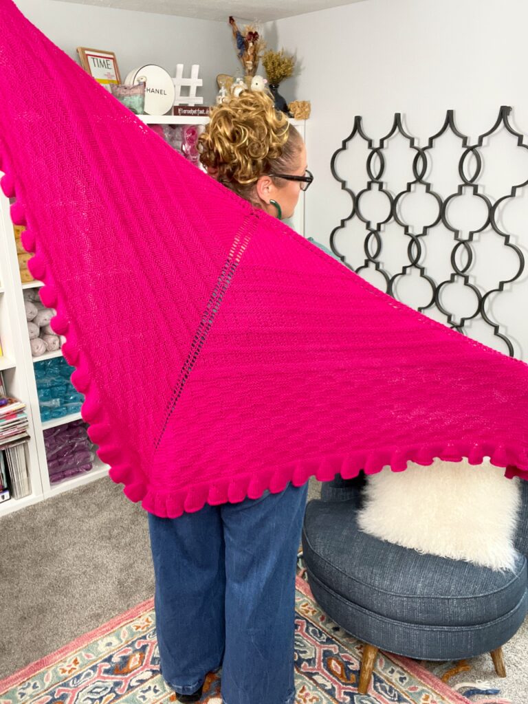 Person displays a bright pink knit shawl with textured edging, highlighting stitch detail in a cozy craft-filled room.