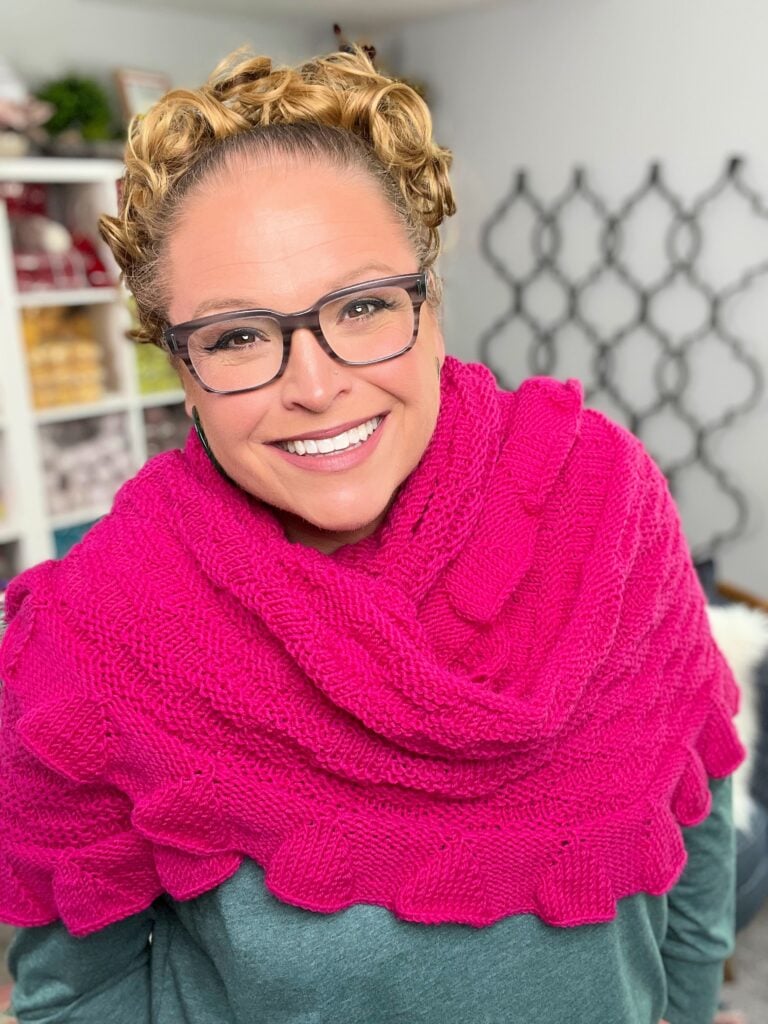 Bright pink knit triangle shawl in worsted yarn, draped over shoulders; visible stitch detail and cozy indoor setting.
