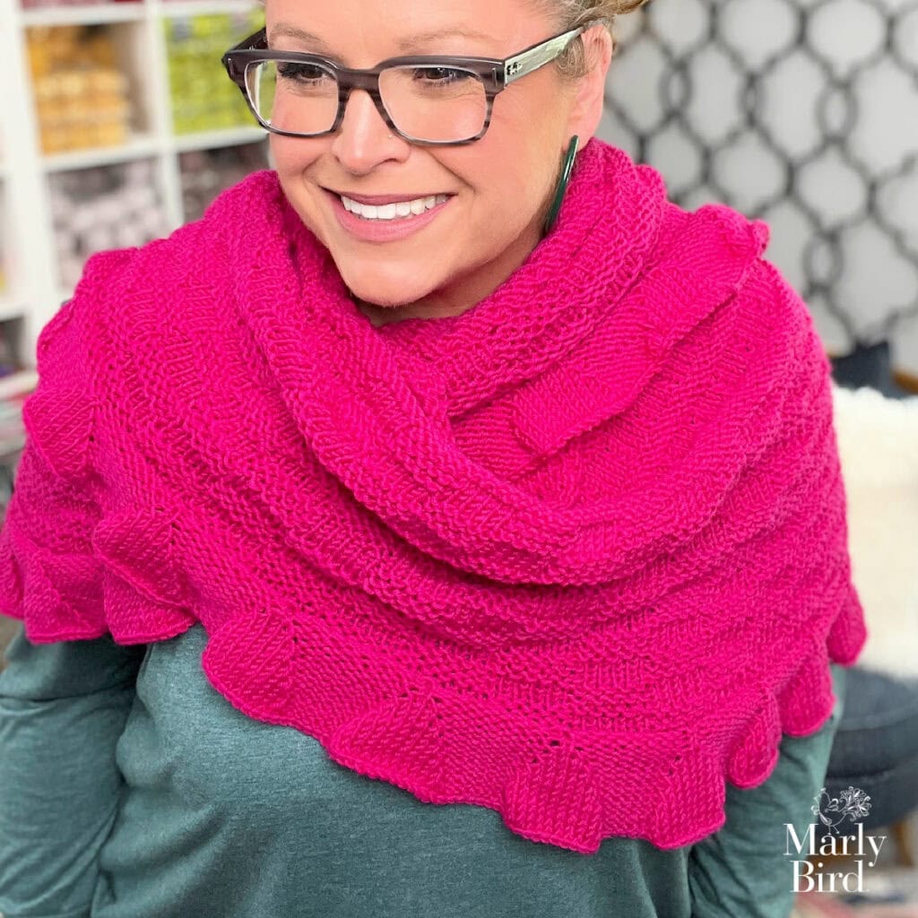 A woman models a bright pink knit shawl with ruffled edges and textured stitches, worn over a green top; yarn shelves behind her.
