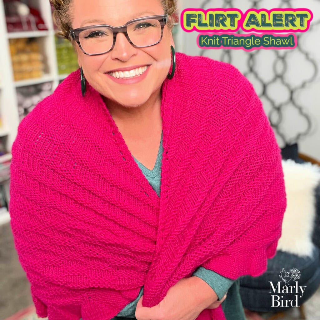 Person wearing a textured, bright pink knit shawl stands in front of bookshelves; "Flirt Alert Knit Triangle Shawl" shown.