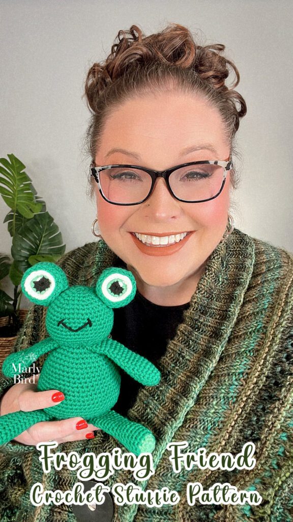 Marly Bird holding the Frogging Friend Crochet Stuffie in one had as she smiles to the camera. The stuffed frog is green with big and bold cartoon eyes. 