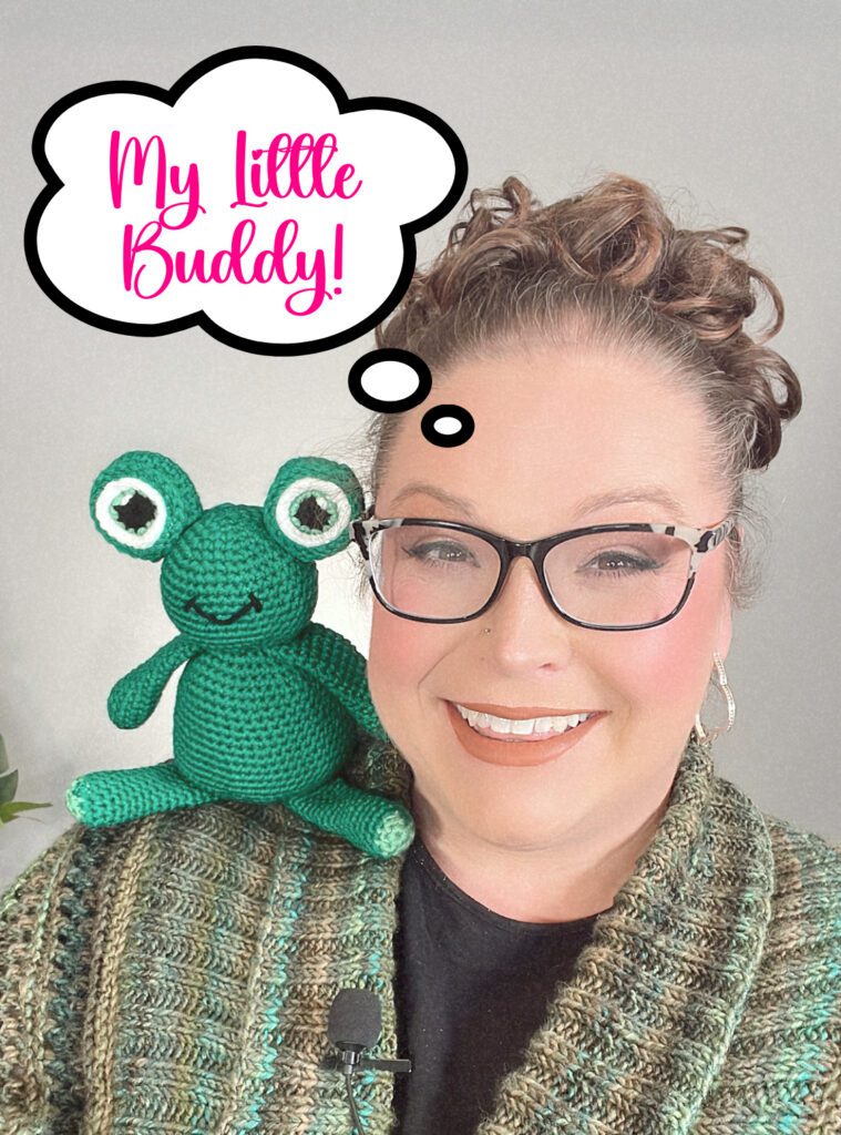 Marly Bird holding the Frogging Friend Crochet Stuffie in one had as she smiles to the camera. The stuffed frog is green with big and bold cartoon eyes.