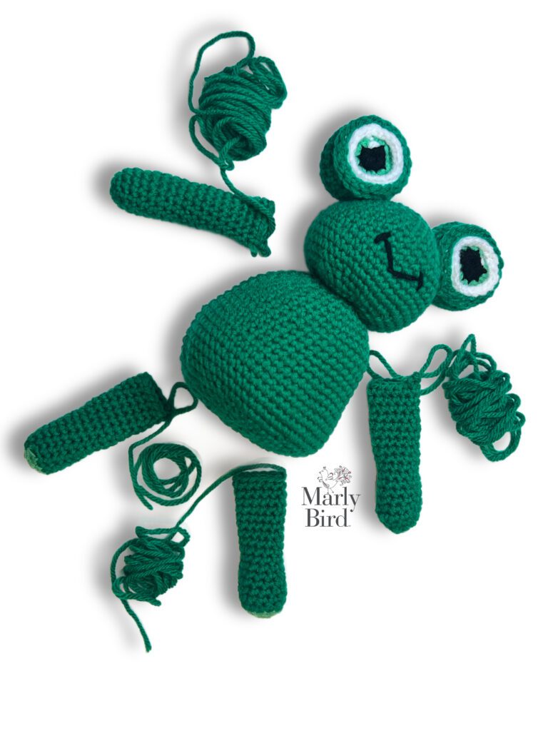 Frogging Friend laying flat on a white surface with the unattached limbs strategically placed along the body. This image demonstrates how the pieces of the stuffed crochet crog should be assembled. This crochet frog pattern is by Marly Bird  