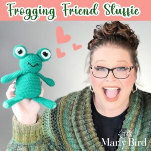 Marly Bird holding the Frogging Friend Crochet Stuffie Pattern in one hand and smiling at the camera. There is text at the top of the image that reads Frogging Friend Stuffie.