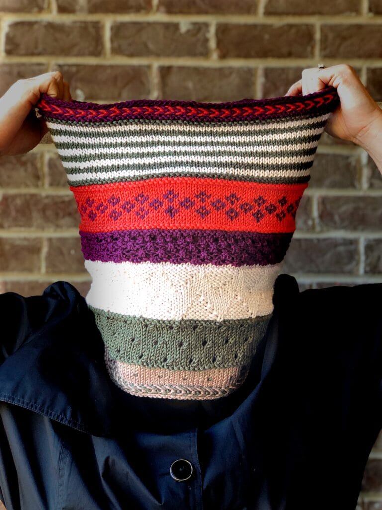 Textured knit cowl with stripes and geometric patterns in multiple colors, shown worn over the head; brick wall backdrop.