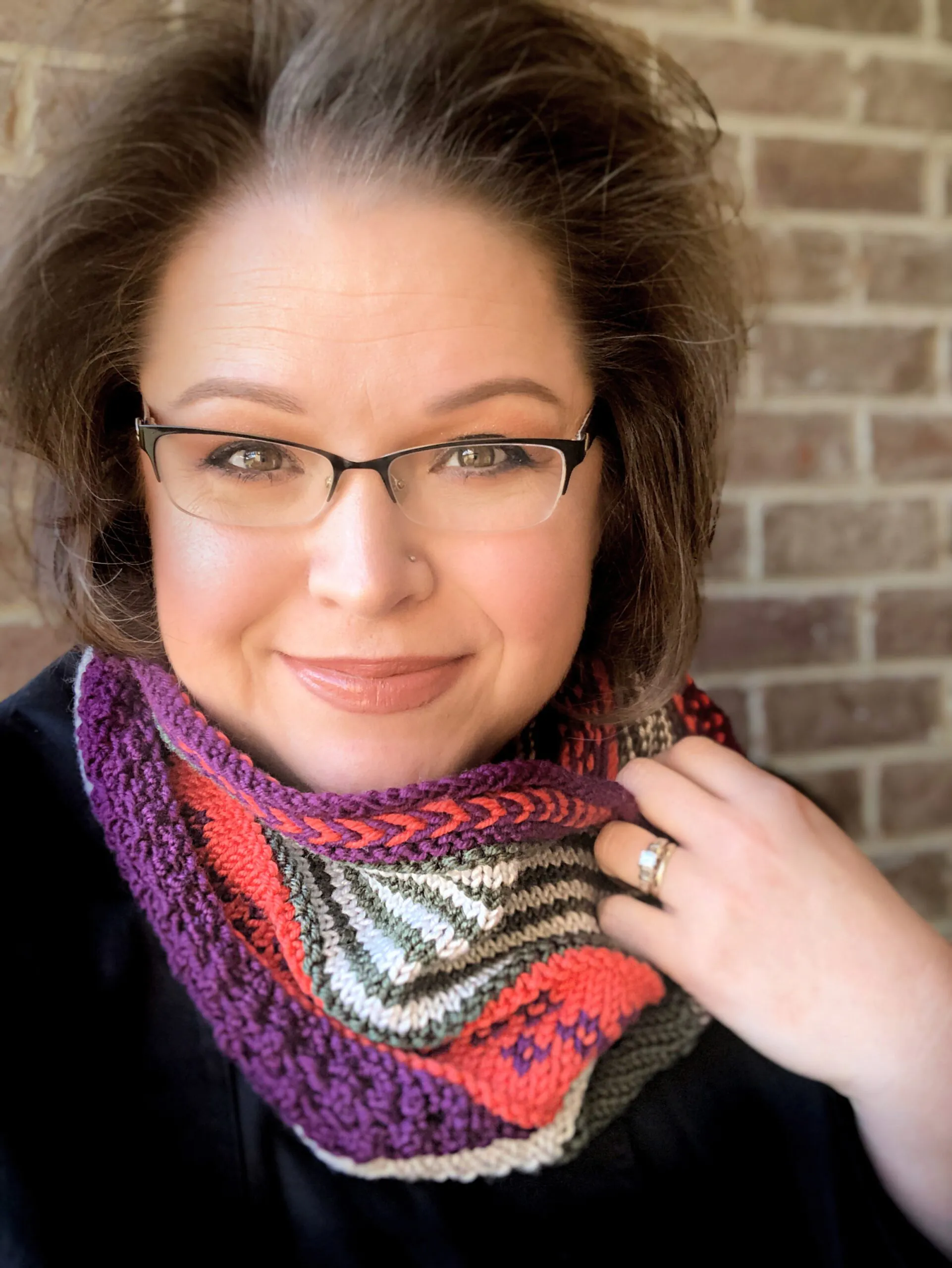 Woman models a colorful knit cowl with visible stitch detail; Marly Bird pattern. Brick wall backdrop. Game Changer knit cowl free pattern with textured stitches