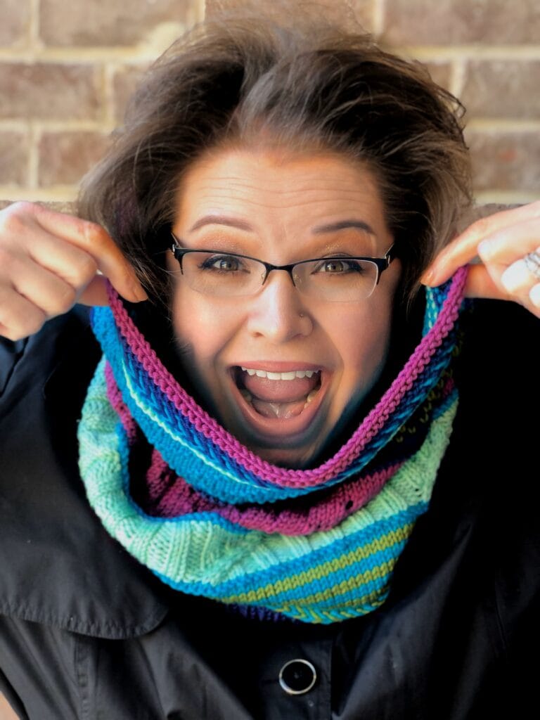 A person models a vibrant knit cowl, showing off its bold colorwork and stitch texture over a black coat, brick wall behind.