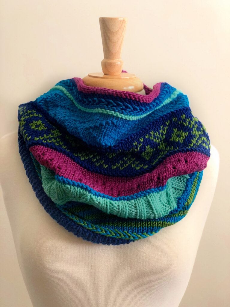 A knit cowl with blue, teal, green, and purple stripes and patterns drapes loosely around a mannequin’s neck.