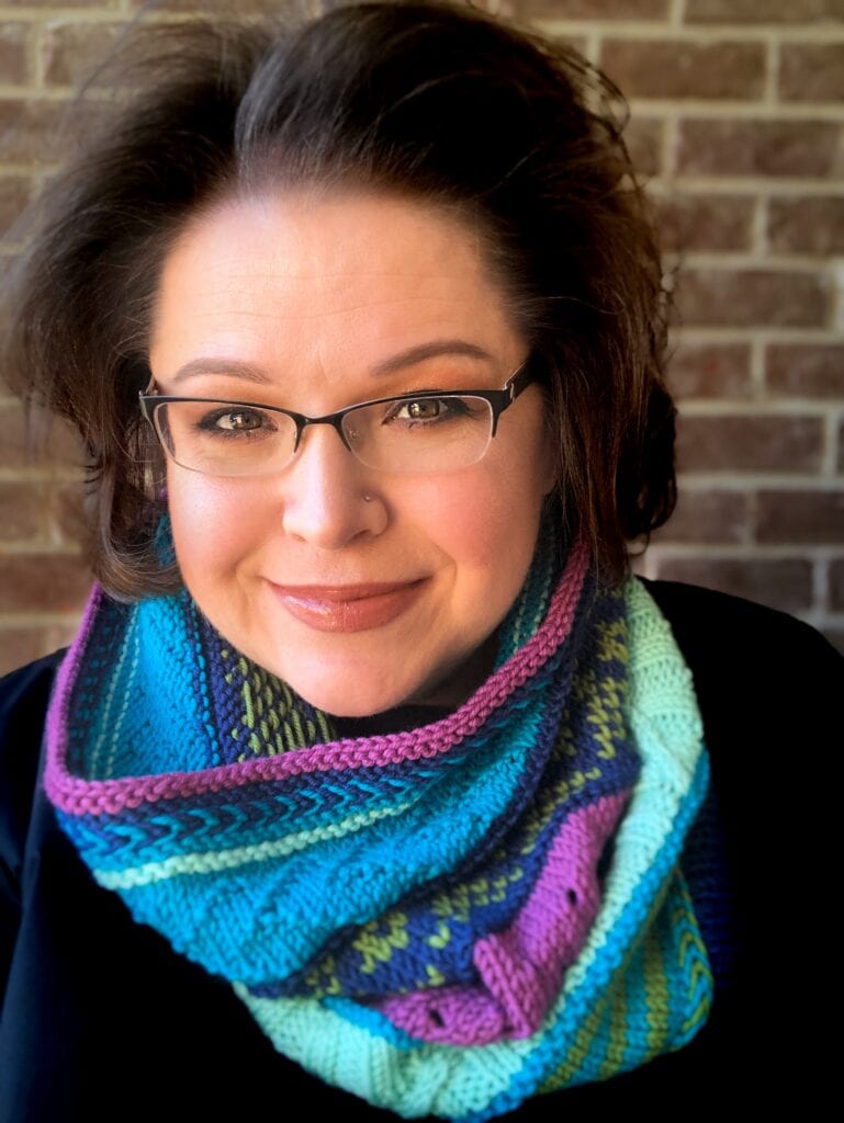 A woman models a knit cowl in blue, purple, and green tones, showing off the cozy texture and stitch details.