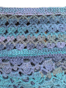 Textured crochet fabric in blue, green, and purple with open shell stitches; intricate for wraps or shawls.