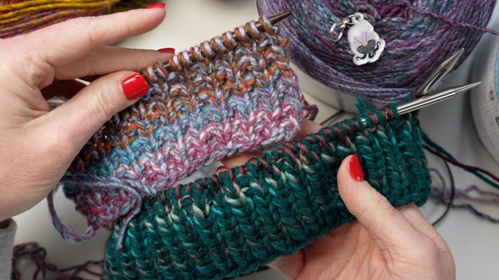 Hands with red nails hold knitting needles working on two textured projects, one in purple-orange, one in green-blue; yarn nearby.