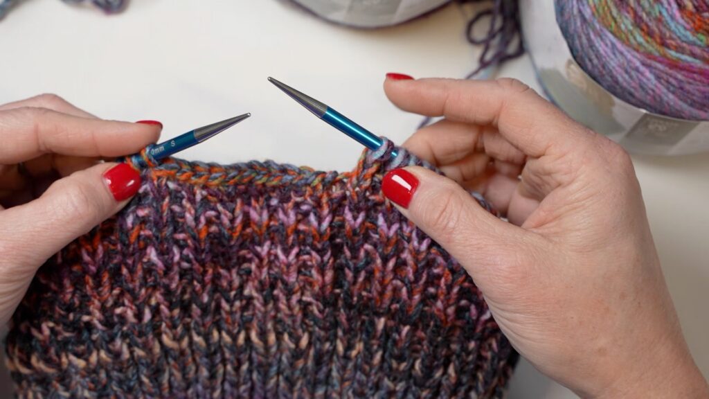 Hands with red polish use blue needles to knit a colorful, textured piece; multicolored yarn balls in the background.