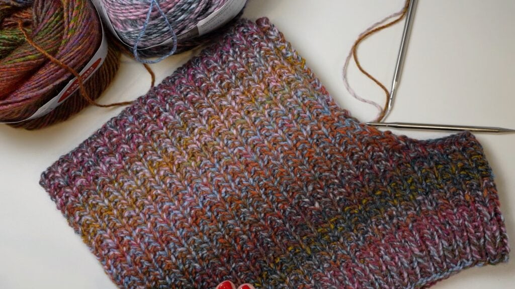 A partially knitted, multicolored fabric with visible stitch detail rests beside two yarn balls and metal needles on a white surface.