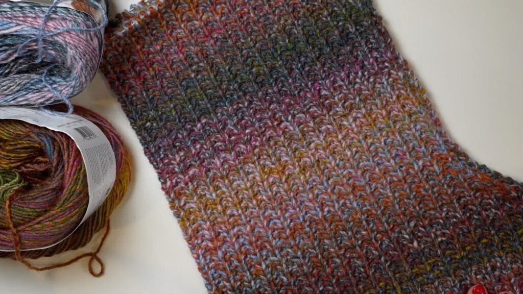 A textured, multicolored scarf in progress rests beside two variegated yarn skeins on a white background.